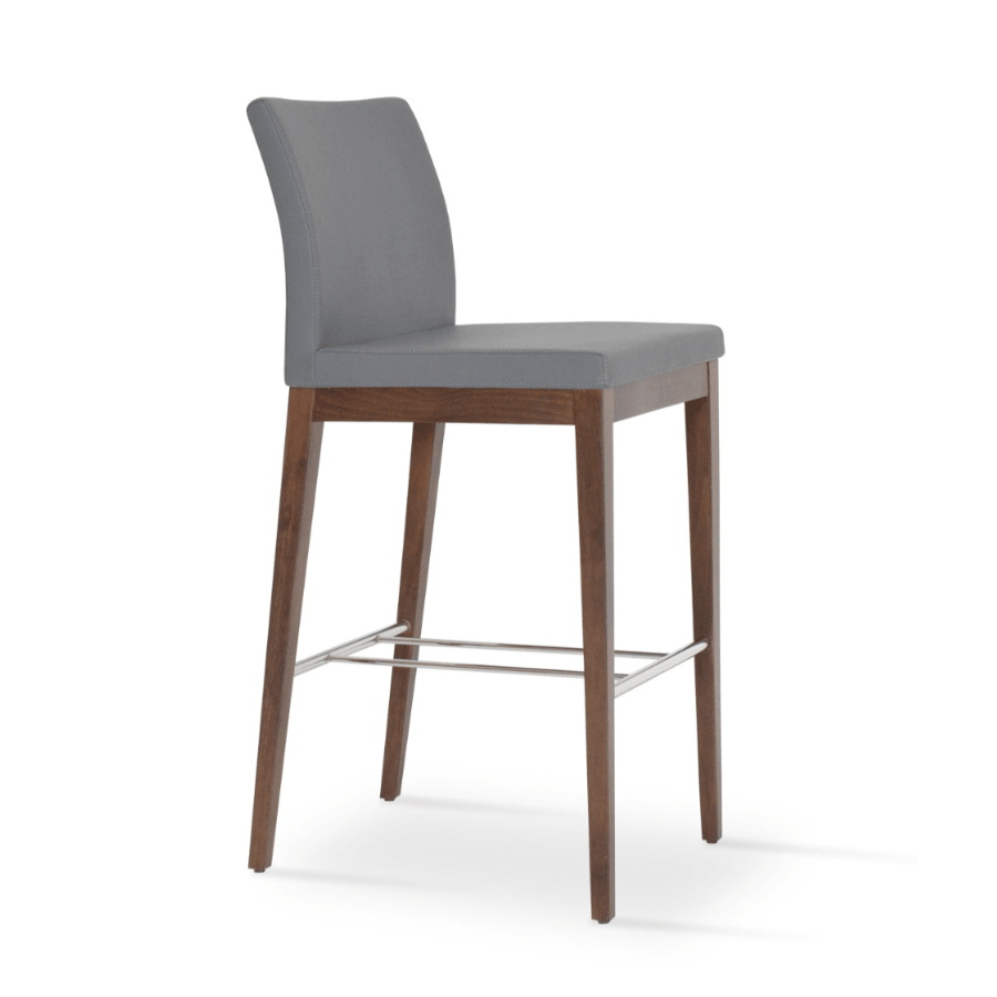 Grey upholstered bar stool with walnut wood legs and chrome footrest against white background